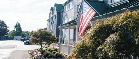 A row of townhouses with a large American flag on the front, a paved driveway, and a small garden with rocks and shrubs.