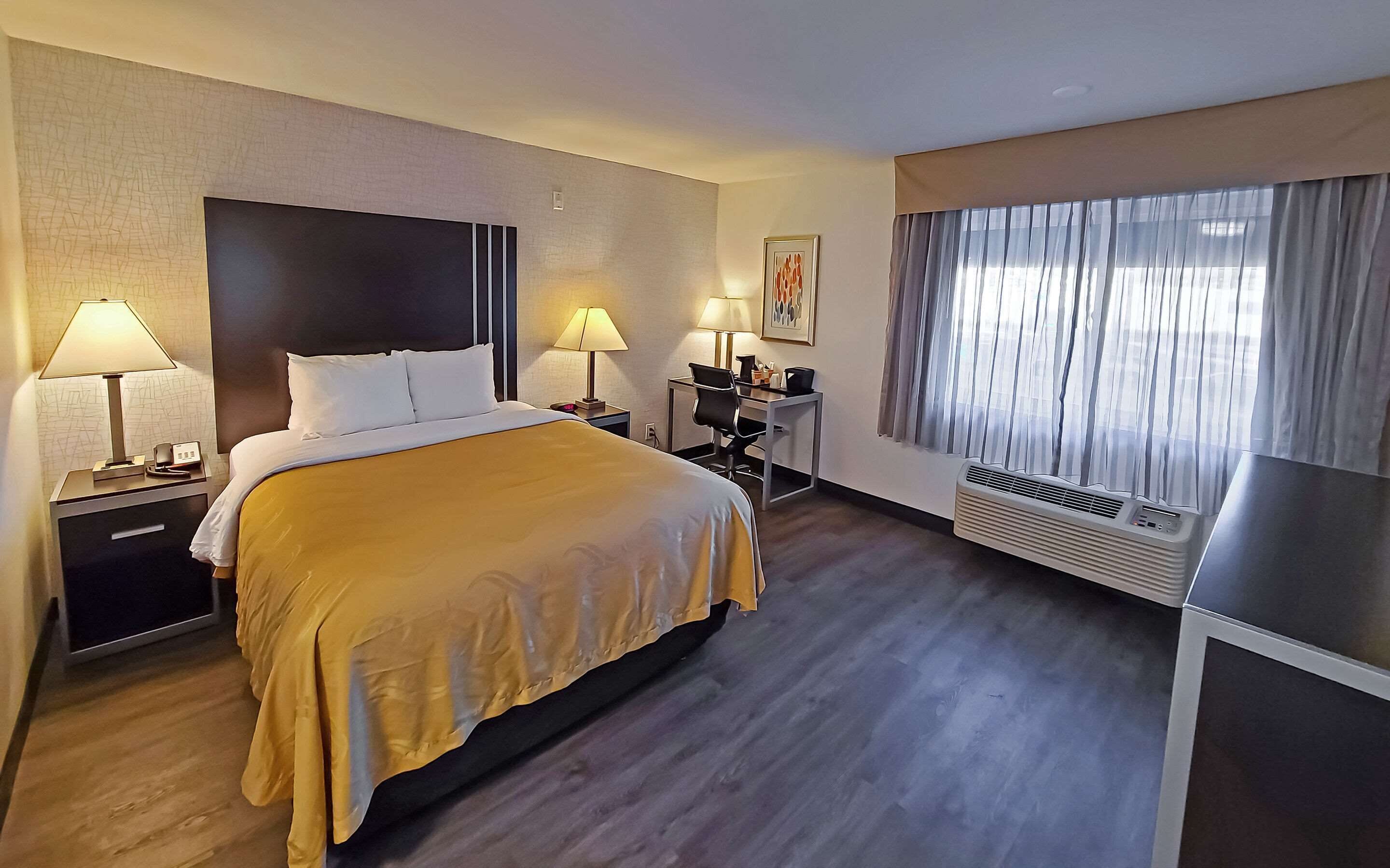 Ein Hotelzimmer mit Bett, Nachttischlampen, einem Schreibtisch mit Stuhl, einem Fenster mit Vorhängen und einer Klimaanlage.