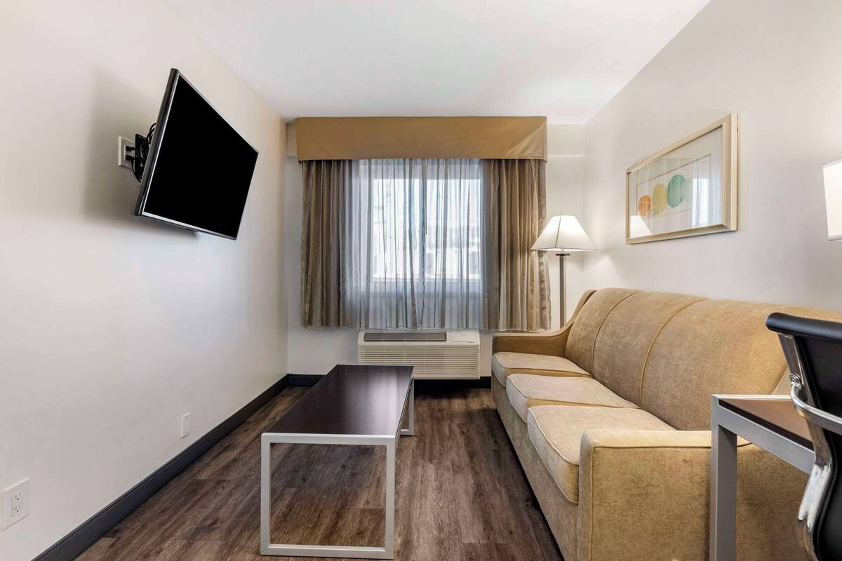 A hotel room with a flat-screen TV mounted on the wall, a sofa, a small table, a lamp, and a window with curtains.