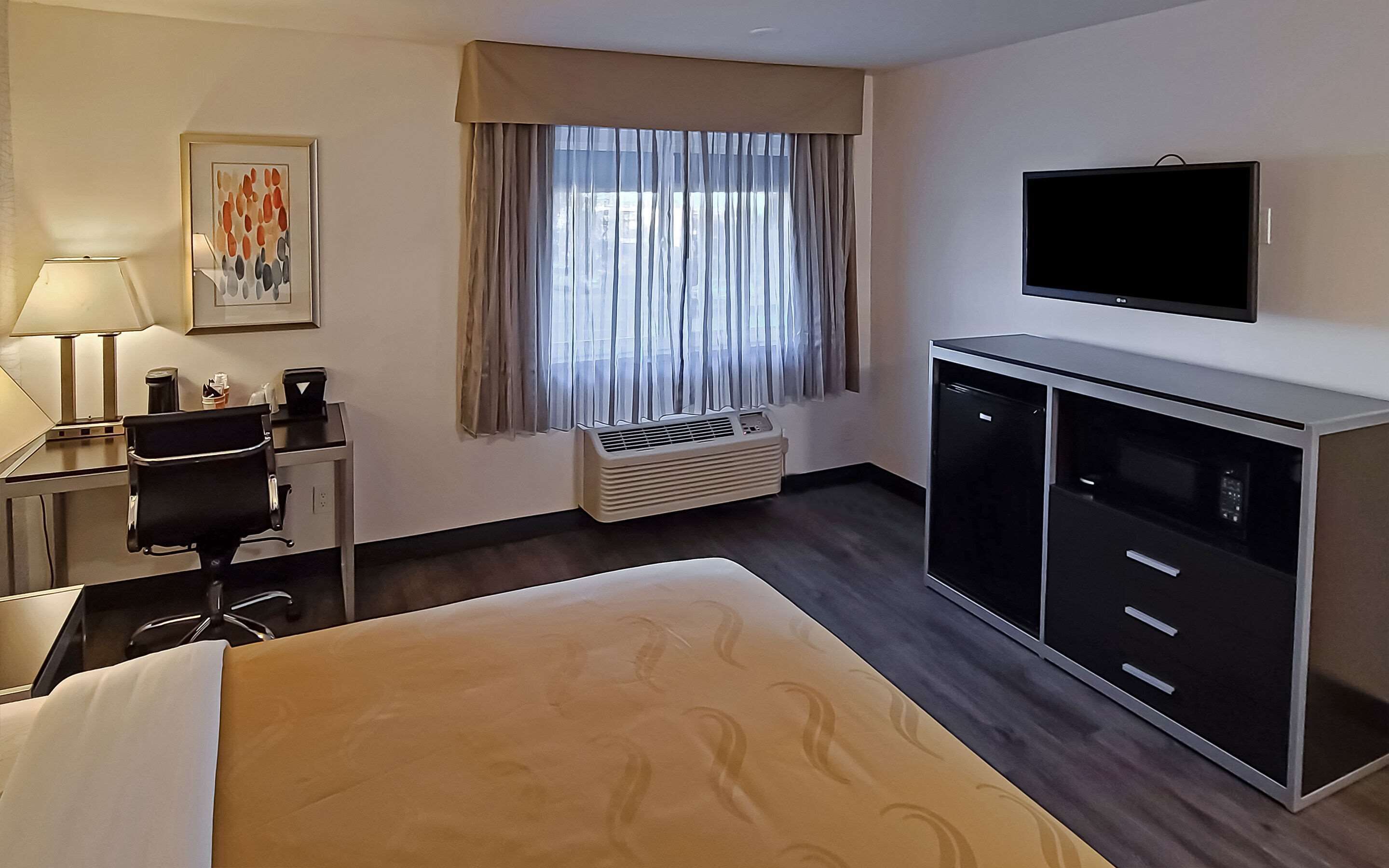 A hotel room with a bed, a desk with a lamp, a television, a minibar, and a window with curtains.