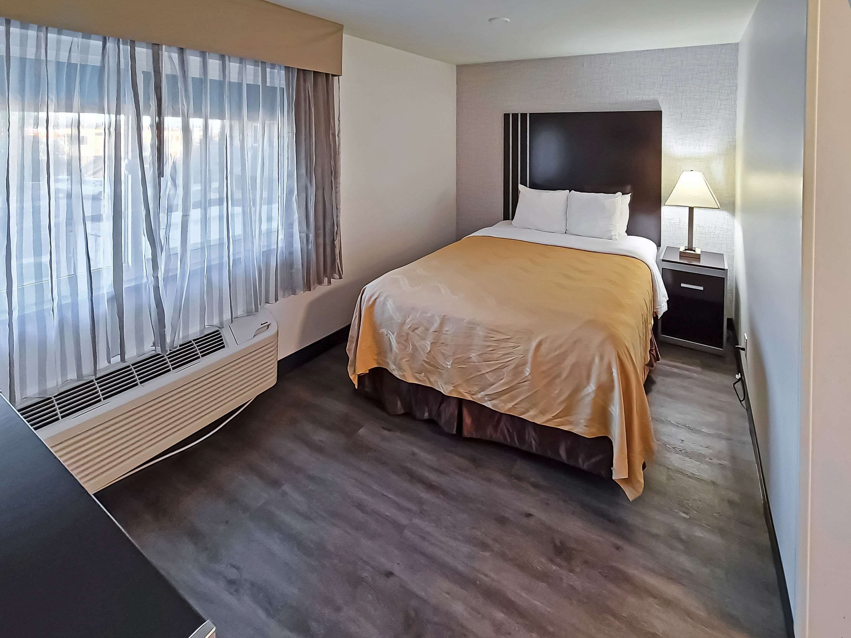 A hotel room with a bed, bedside table, lamp, air conditioning unit, and a window with curtains.