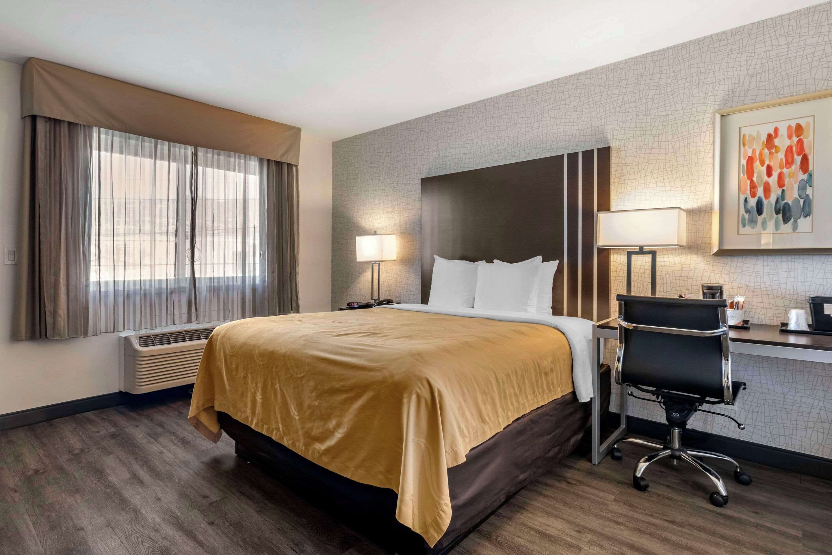 Een hotelkamer met een groot bed, een bureau met stoel, een raam met gordijnen, een wandlamp en een kunstwerk in een lijst.