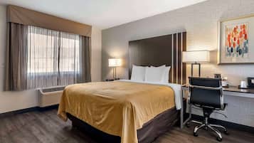 Een hotelkamer met een groot bed, een bureau met stoel, een raam met gordijnen, een wandlamp en een kunstwerk in een lijst.