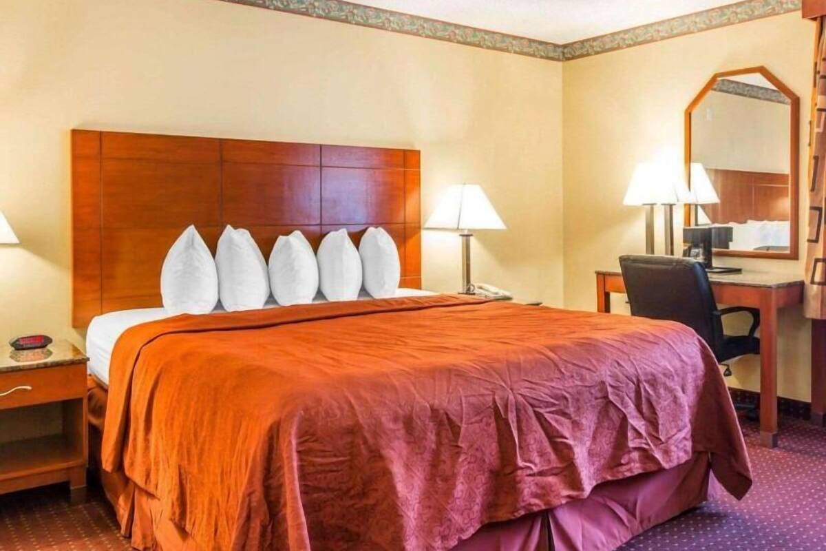 A hotel room with a wooden headboard, a large bed with a red bedspread, two bedside lamps, a desk with a chair, and a mirror on the wall.