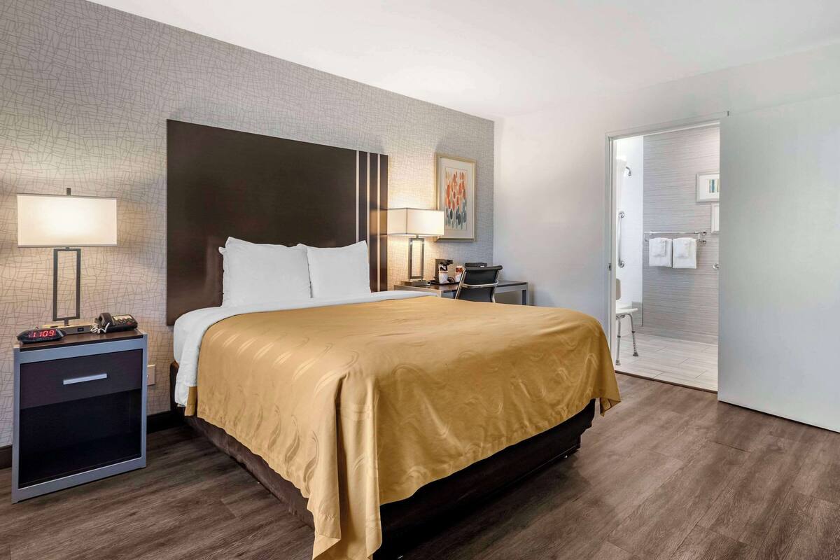 A hotel room with a bed, bedside tables, a nightstand, a lamp, a painting, and a bathroom visible through an open door.