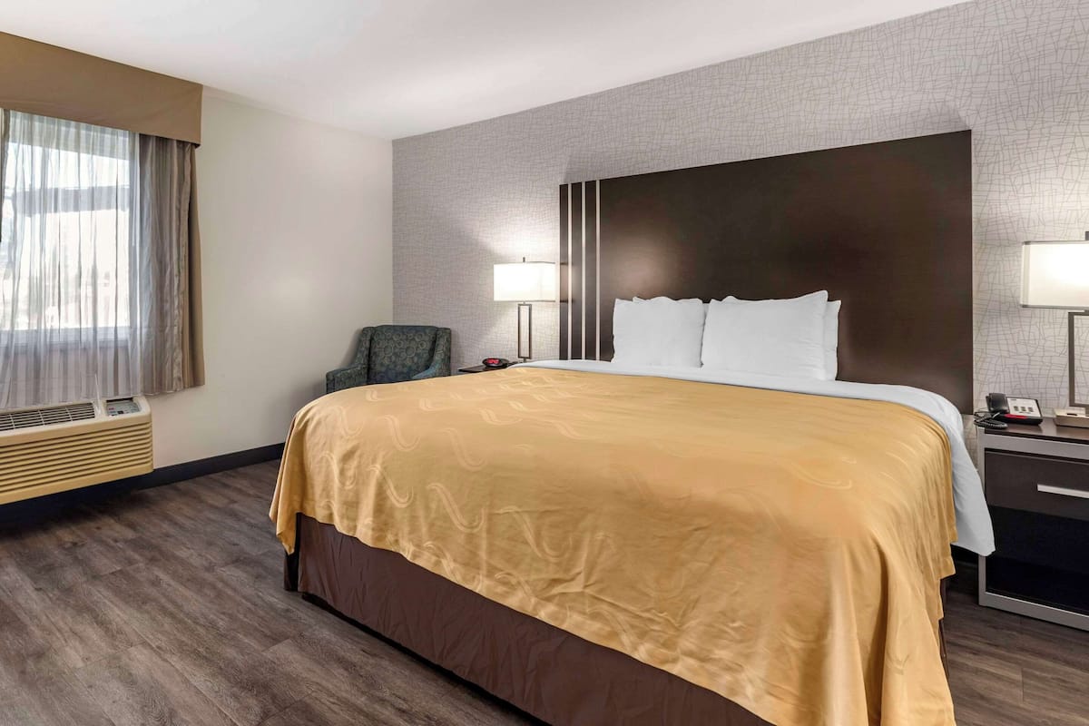 A hotel room with a large bed, a chair, a bedside table with a lamp, and a wall-mounted air conditioner.