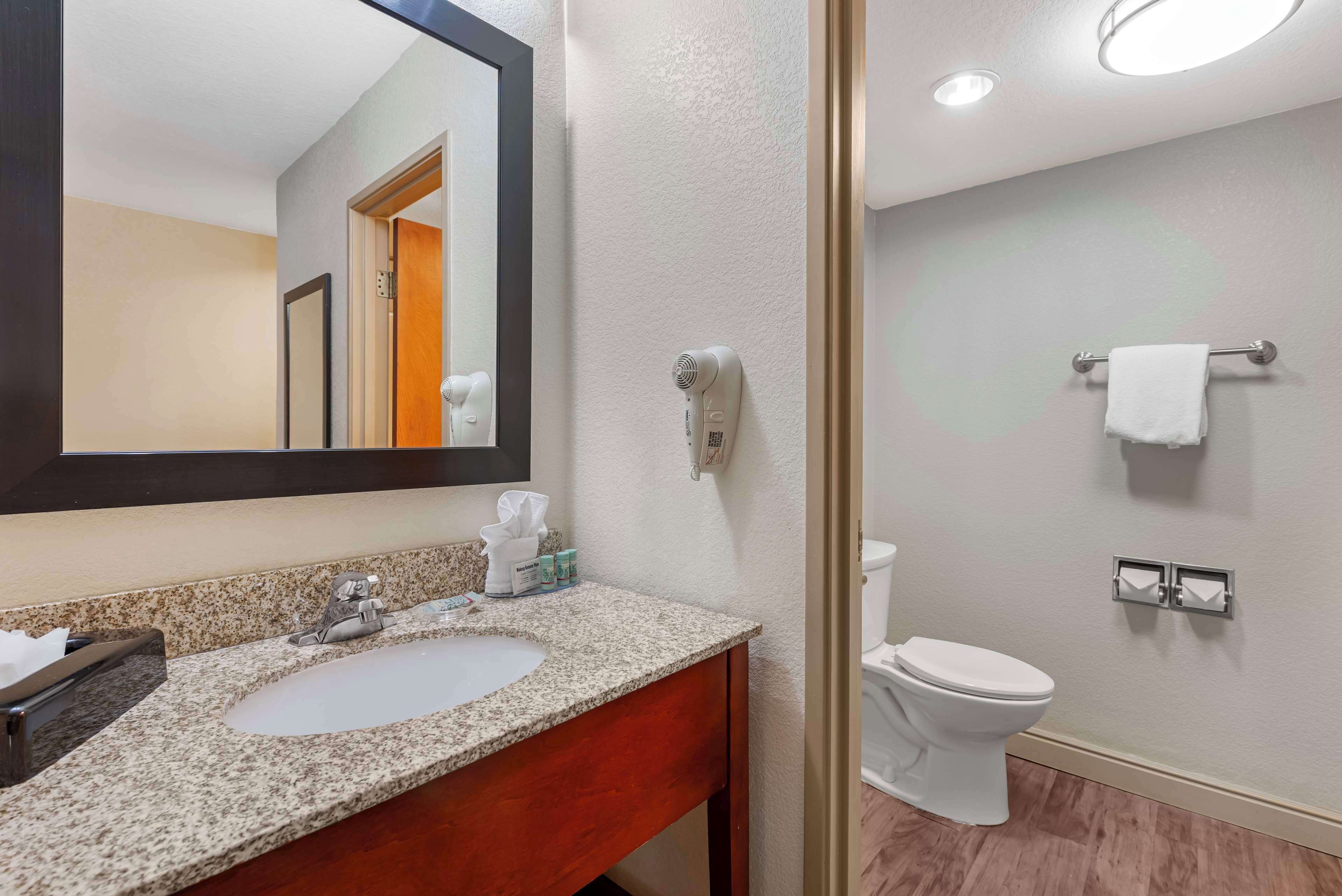 A bathroom with a granite countertop sink, a mirror, a towel rack, a toilet, and a hairdryer mounted on the wall.