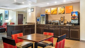 A hotel dining area with a wooden table and red upholstered chairs, a coffee machine, and a juice dispenser. - Comfort Inn & Suites Lake Norman (Cornelius)
