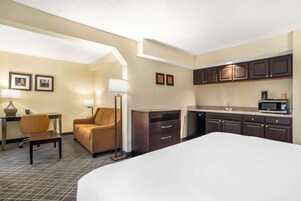 Habitación de hotel con cama, escritorio con silla, sofá, kitchenette con microondas y mesita de noche con lámpara.