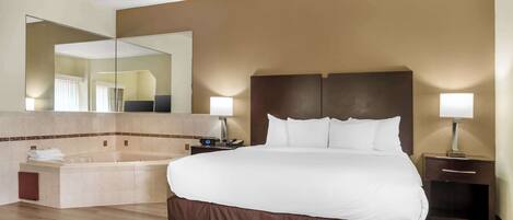A hotel room with a large bed, a built-in bathtub, a bedside table with a lamp, and a large mirror above the bed.
