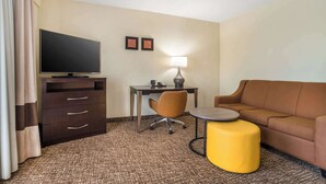 A hotel room with a flat-screen TV, a desk with a lamp, a brown leather sofa, a yellow ottoman, and a patterned carpet. - Comfort Inn & Suites Lake Norman (Cornelius)
