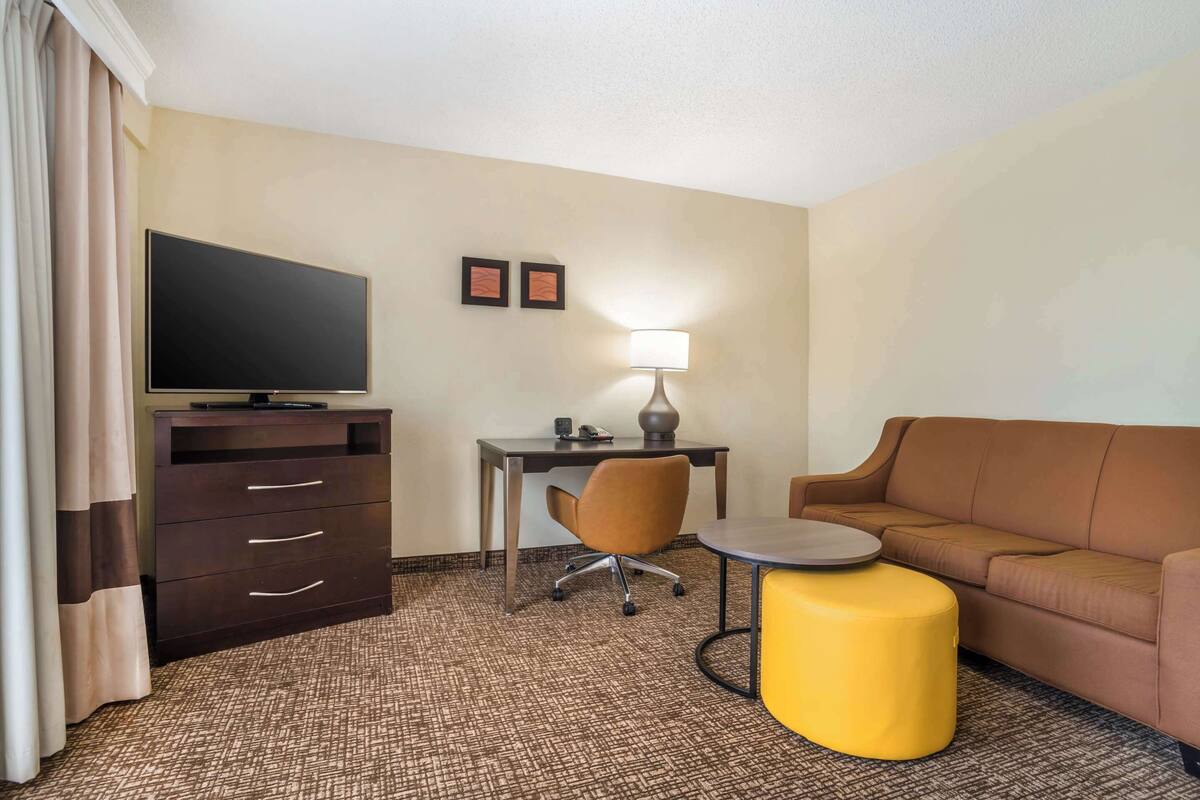 A hotel room with a flat-screen TV, a desk with a lamp, a brown leather sofa, a yellow ottoman, and a patterned carpet.