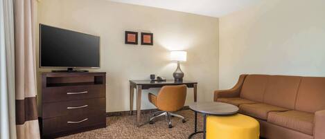 Habitación de hotel con televisor de pantalla plana, escritorio con lámpara, sofá de cuero marrón, reposapiés amarillo y alfombra estampada.