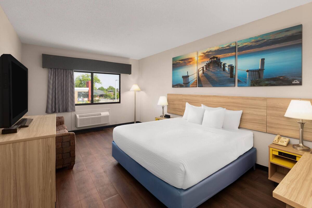 A hotel room with a large bed, a desk, a TV, a window with curtains, a lamp, and a painting on the wall.