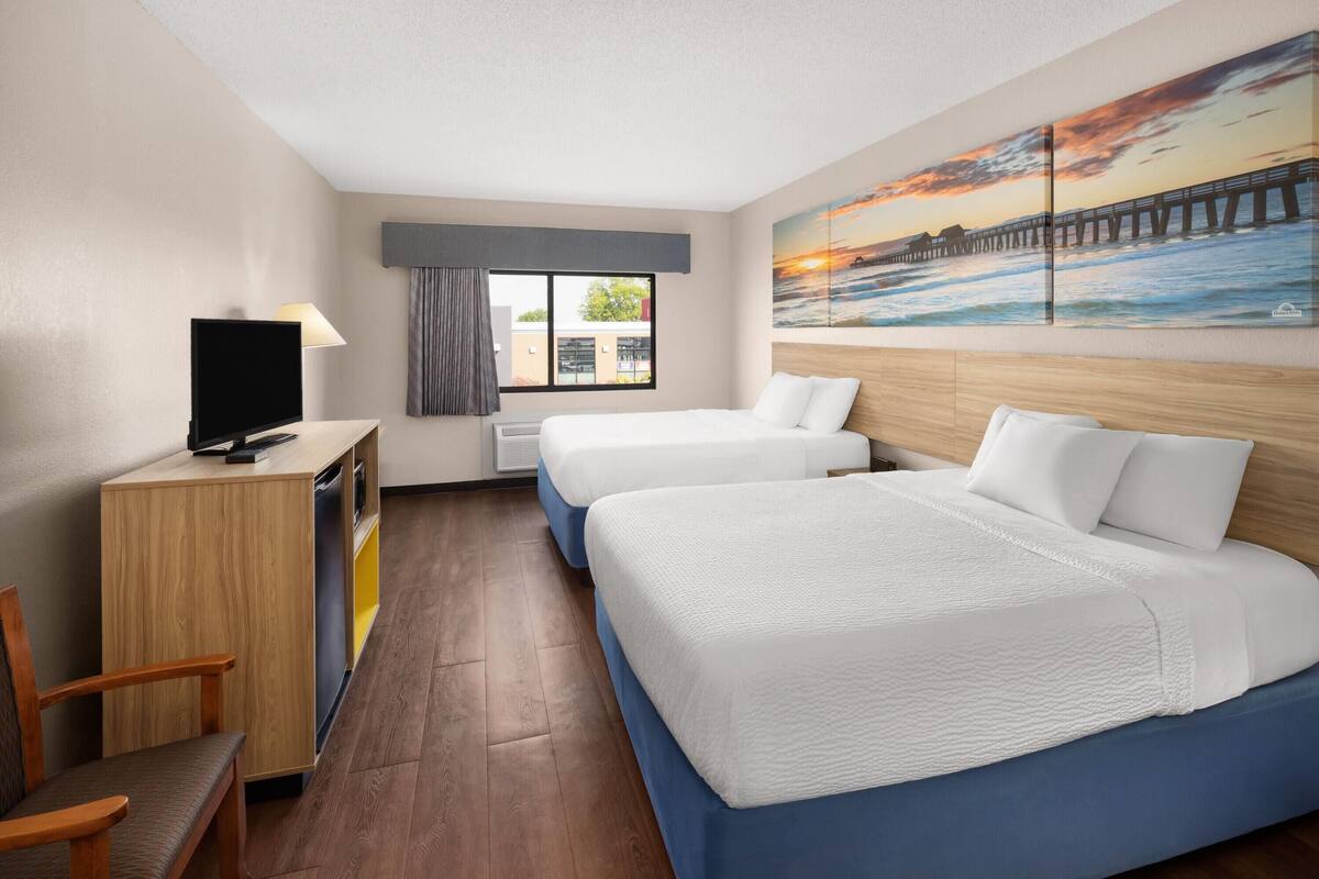 A hotel room with two beds, a desk with a TV, a window with curtains, and a painting of a sunset over a pier.