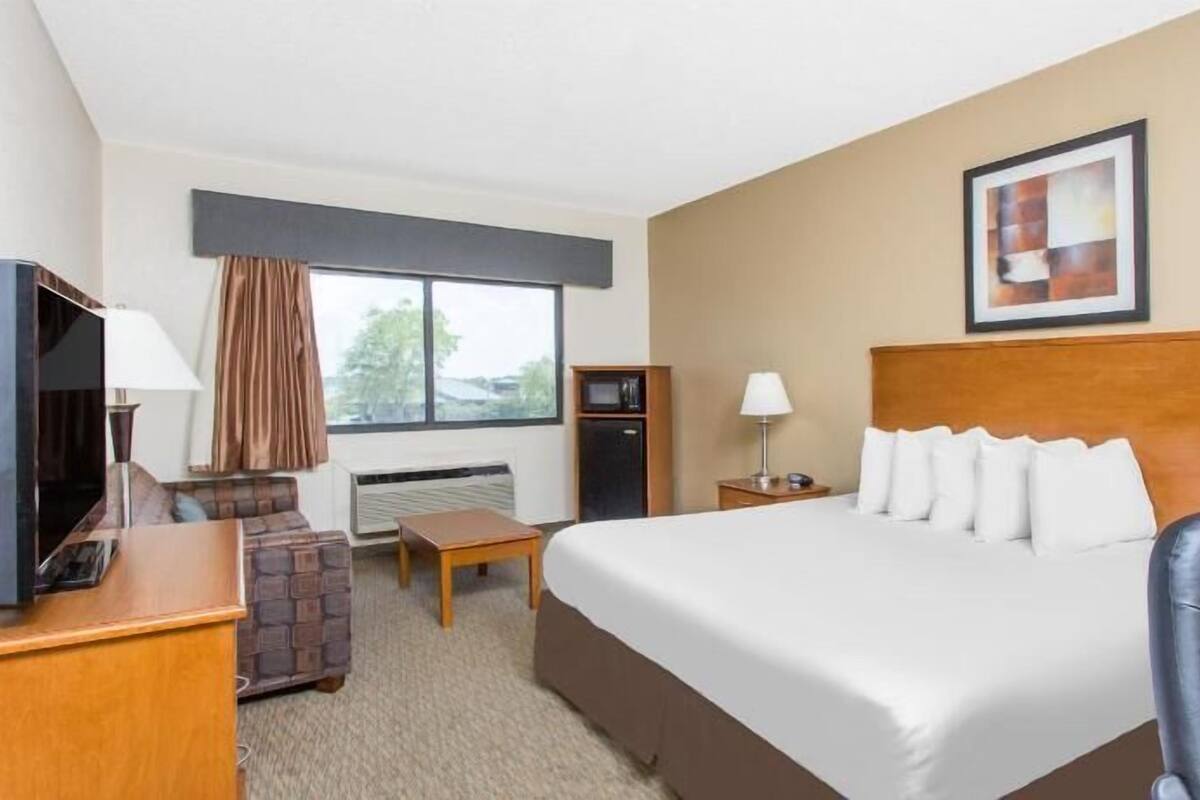 A hotel room with a large bed, a desk with a TV, a chair, a small table, and a window with curtains.