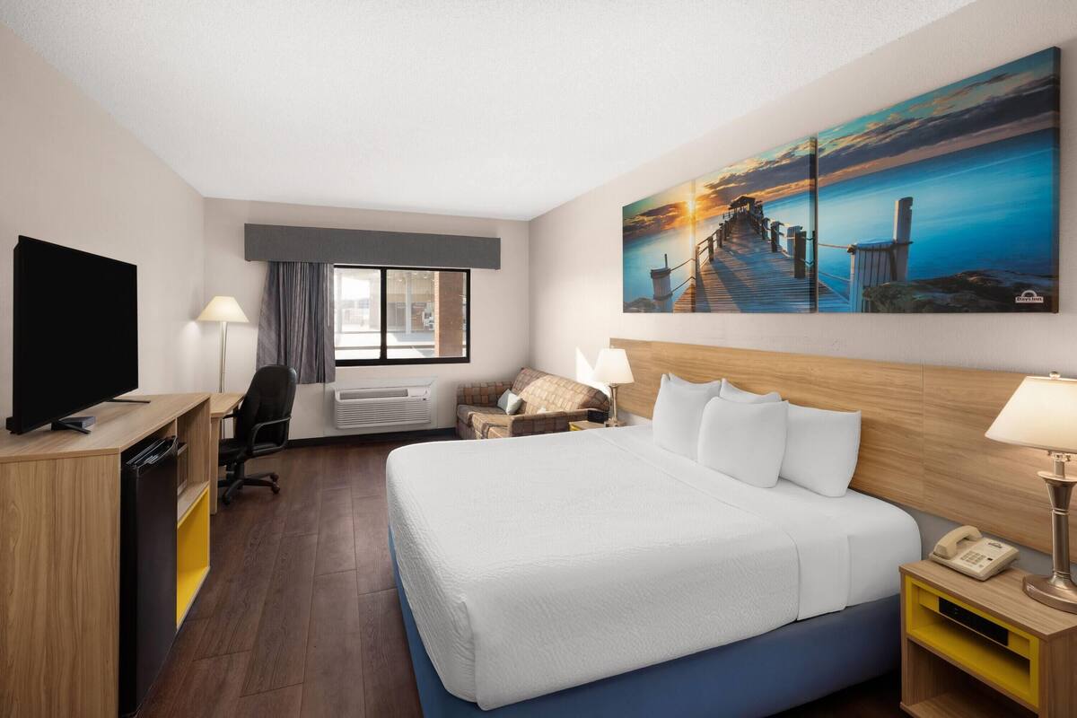 A hotel room with a large bed, a desk with a TV, a chair, a window with curtains, a painting of a sunset, and a nightstand with a lamp.