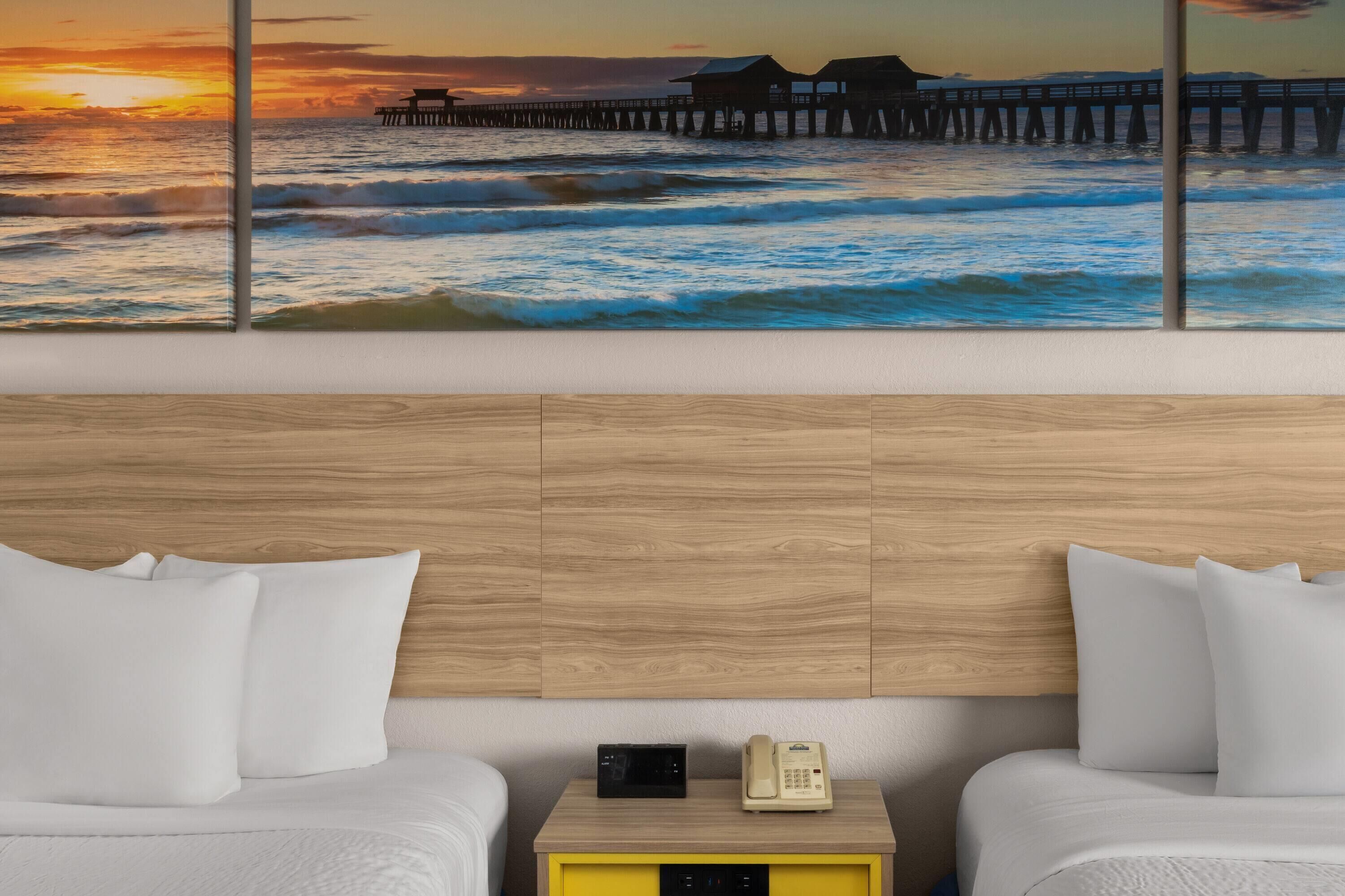 A hotel room with a wooden headboard, two beds with white bedding, a nightstand with a phone and a digital clock, and a large ocean view painting.