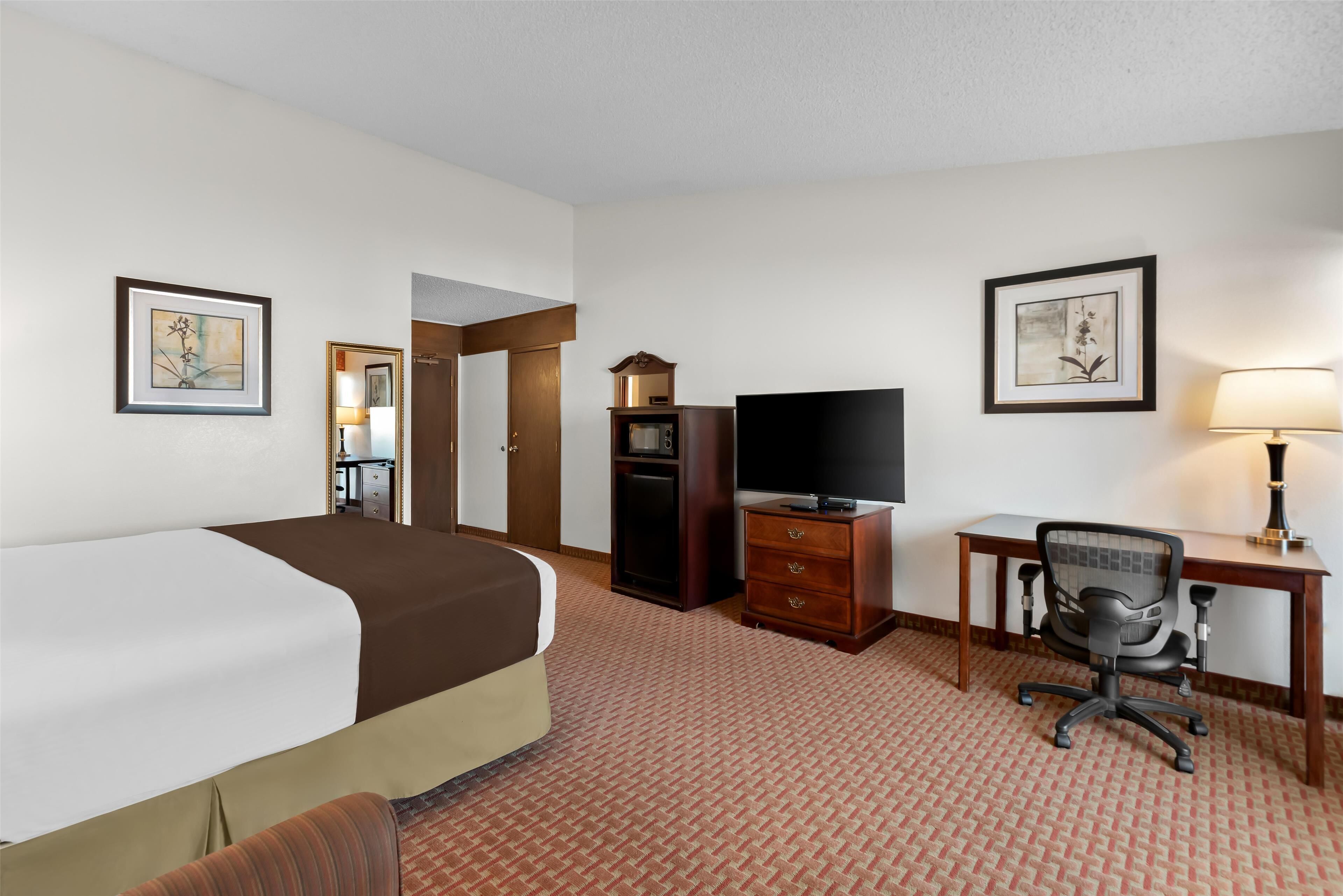 A hotel room with a bed, a desk with a chair, a television, a dresser, and framed artwork on the walls.