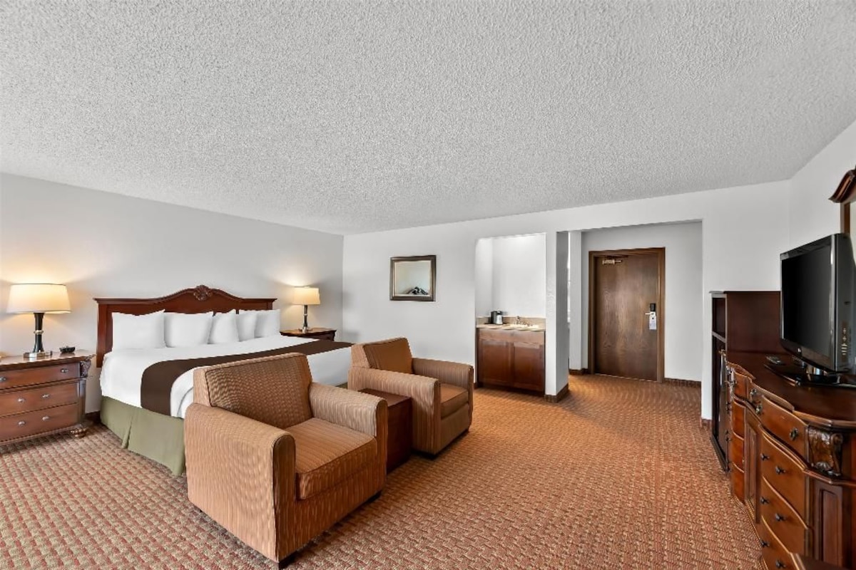 A hotel room with a bed, two armchairs, a dresser, a television, and a kitchenette.