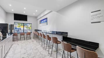 A modern bar area with marble flooring, a long counter, and several cushioned bar stools. A sign indicates complimentary breakfast hours.