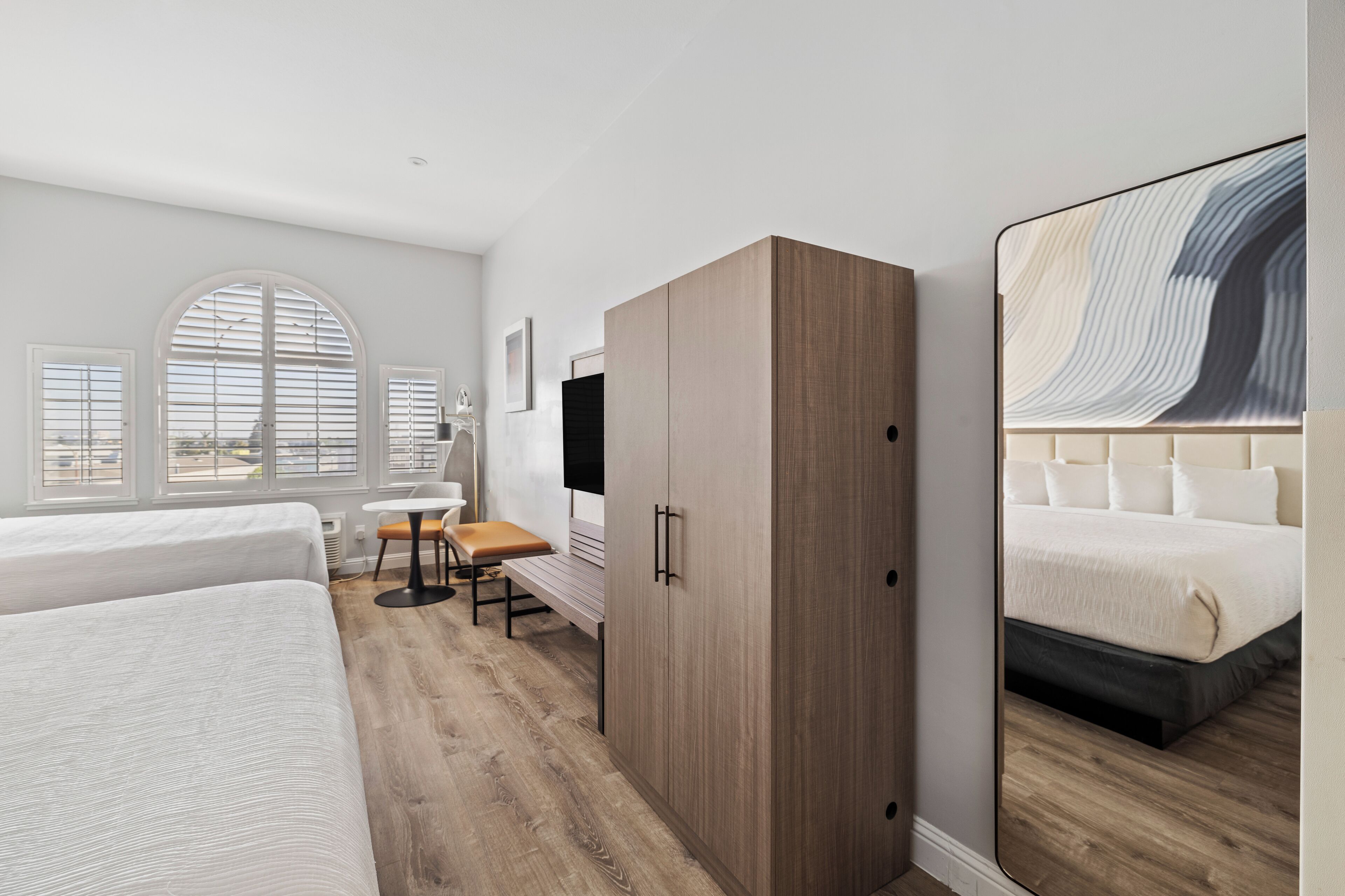 A modern hotel room with a large bed, a wooden wardrobe, a desk with a chair, and a large mirror reflecting the bed.