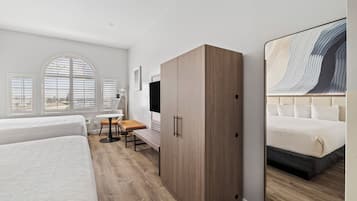 A modern hotel room with a large bed, a wooden wardrobe, a desk with a chair, and a large mirror reflecting the bed.