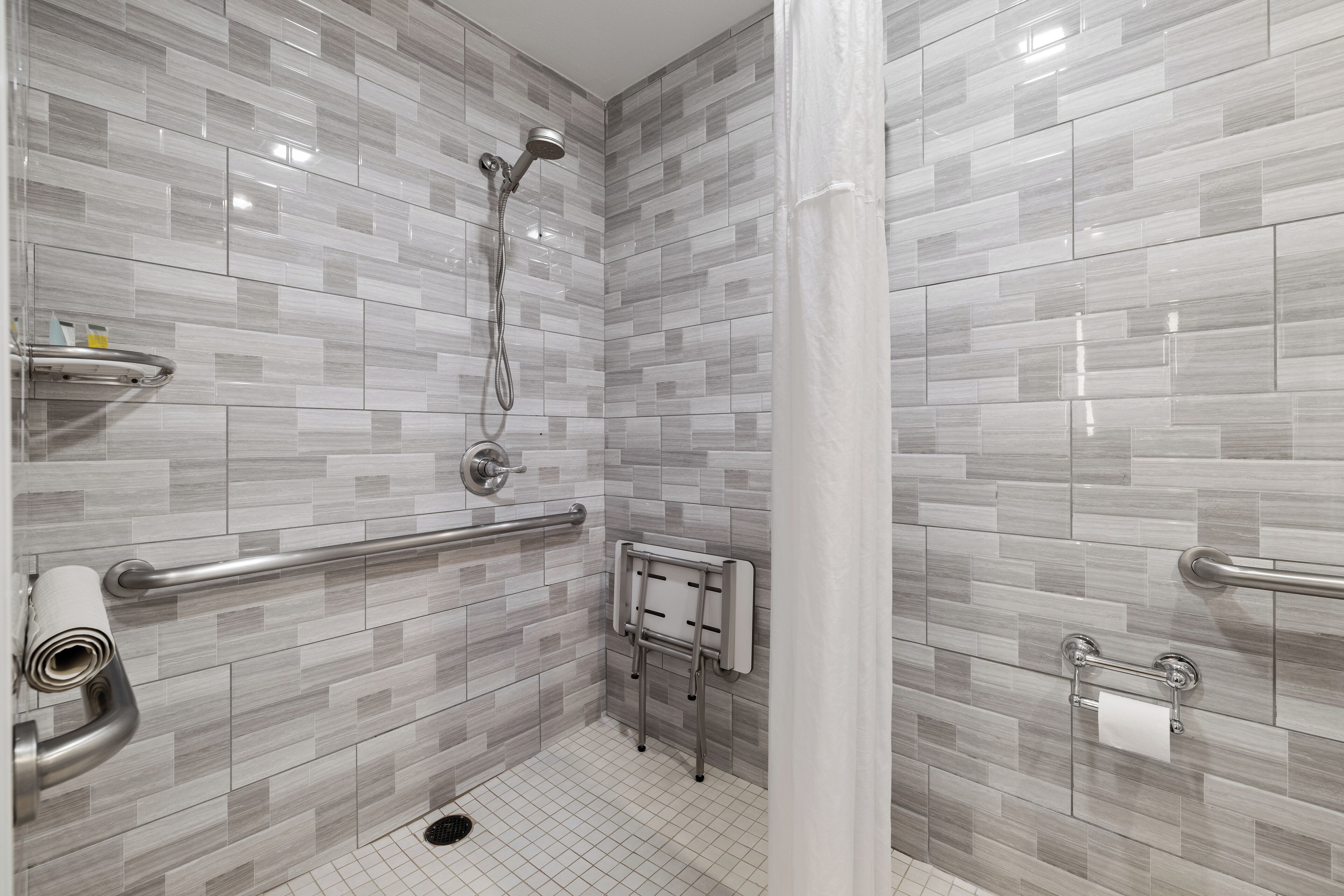 Um banheiro moderno com azulejos cinzas e brancos, chuveiro com chuveiro manual, barra de apoio, porta-toalhas e suporte para papel higiênico.