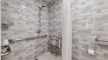 Um banheiro moderno com azulejos cinzas e brancos, chuveiro com chuveiro manual, barra de apoio, porta-toalhas e suporte para papel higiênico.