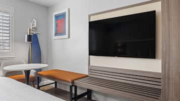 A modern hotel room with a large flat-screen TV mounted on the wall, a small round table with a bench, and a ceiling fan.