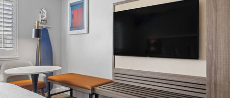 A modern hotel room with a large flat-screen TV mounted on the wall, a small round table with a bench, and a ceiling fan.