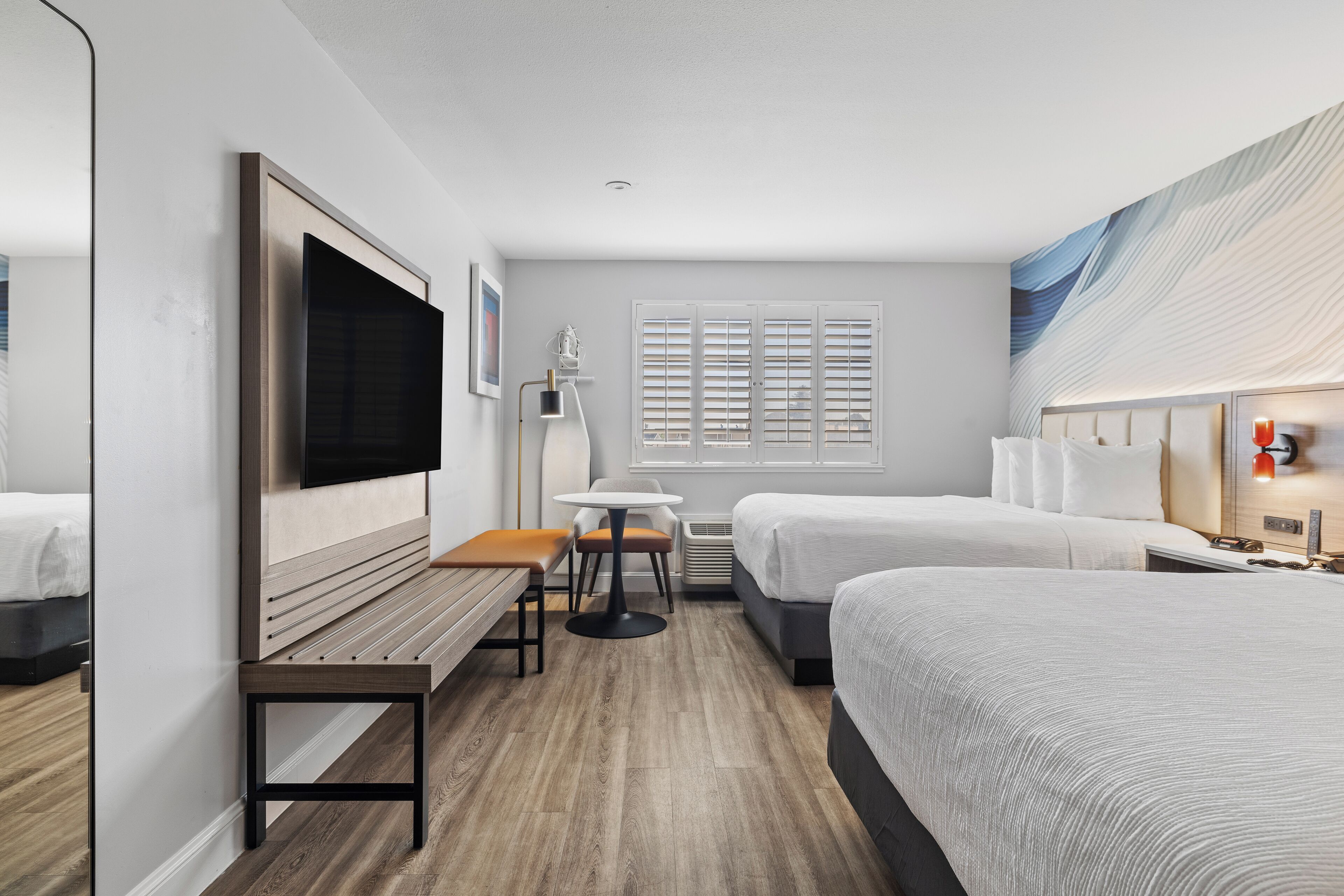 Una habitación de hotel con una cama grande, una cama más pequeña, un escritorio con silla, televisor, ventana con persianas y una lámpara fijada a la pared.