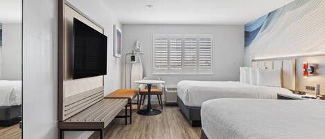 Una habitación de hotel con una cama grande, una cama más pequeña, un escritorio con silla, televisor, ventana con persianas y una lámpara fijada a la pared.