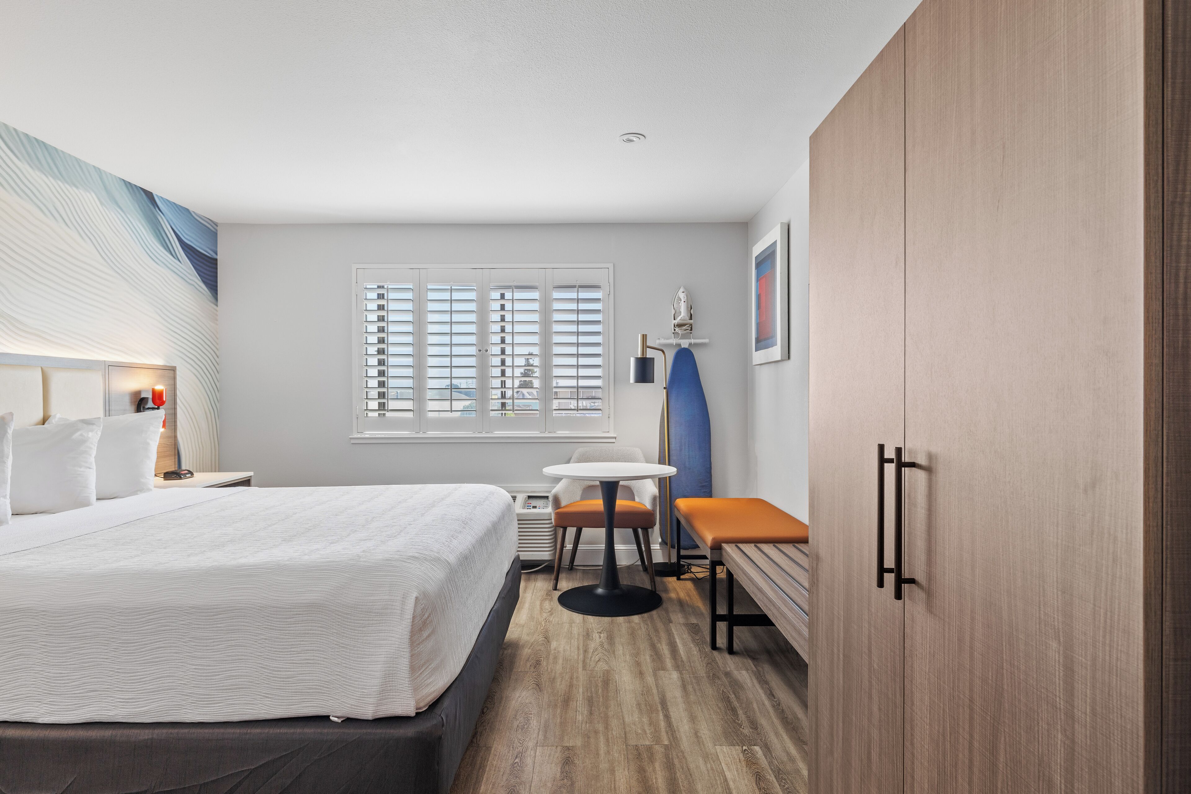 A modern hotel room with a large bed, a small round table, a bench, and a wardrobe.