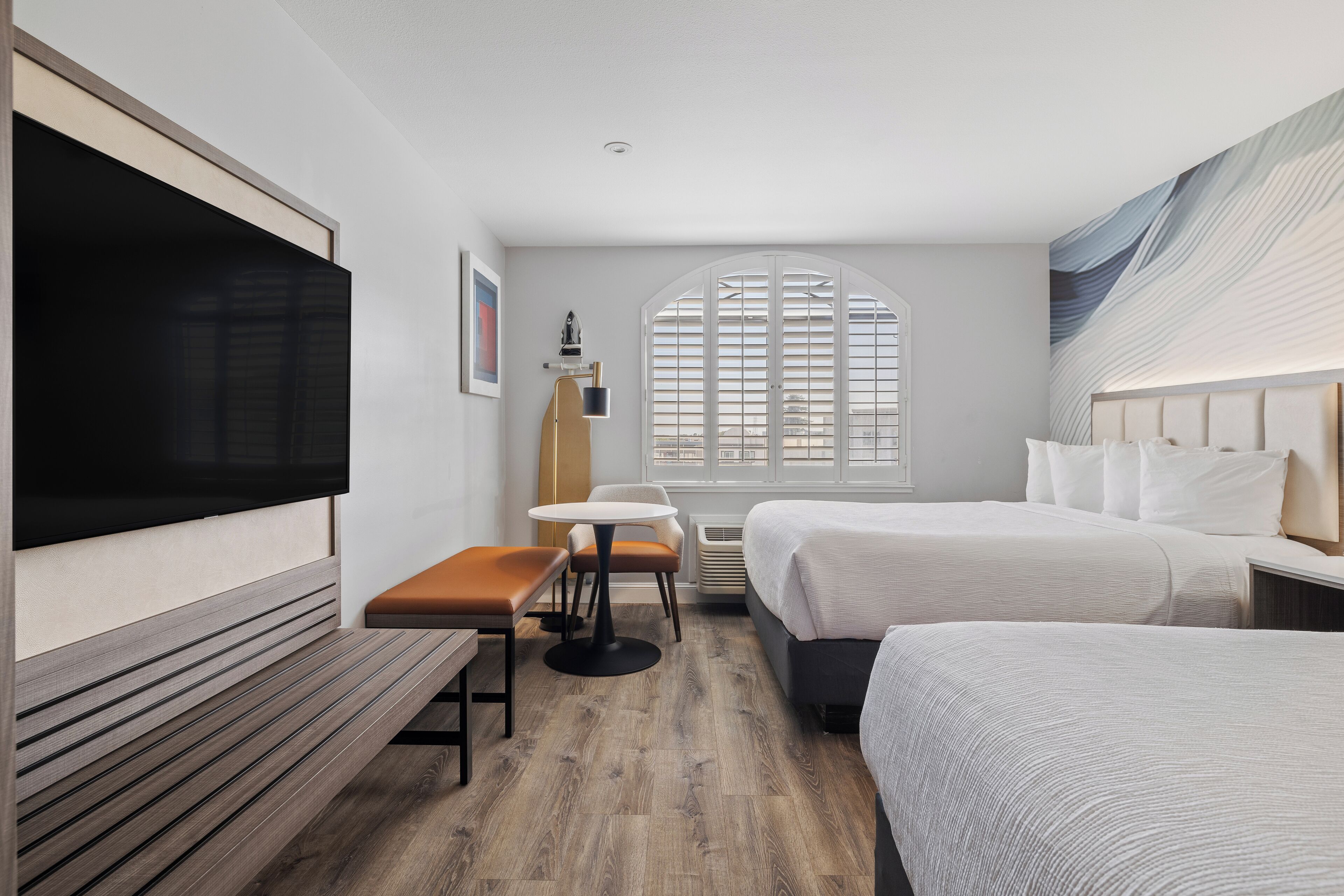 A hotel room with a large flat-screen TV, a wooden bench, a small round table, two beds, and a large window with blinds.