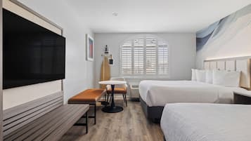 A hotel room with a large flat-screen TV, a wooden bench, a small round table, two beds, and a large window with blinds.