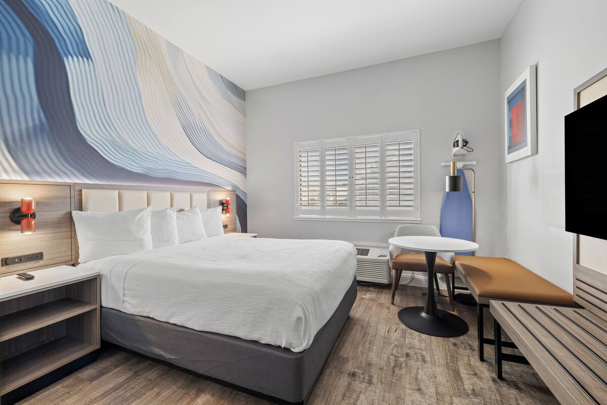 A modern hotel room with a large bed, bedside tables, a desk, a chair, a small table, and a TV.