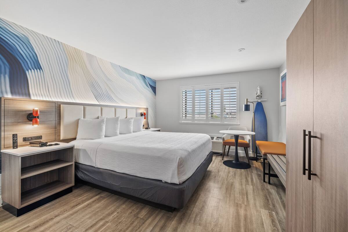 A modern hotel room with a large bed, a desk, and a chair. There is a window with blinds, a wall-mounted lamp, and a decorative wall piece.
