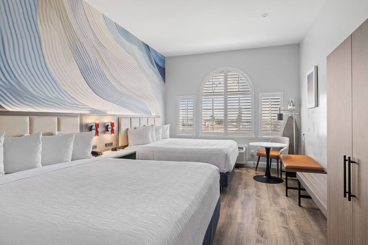 A hotel room with two beds, a large window with blinds, a small table, and a bench.