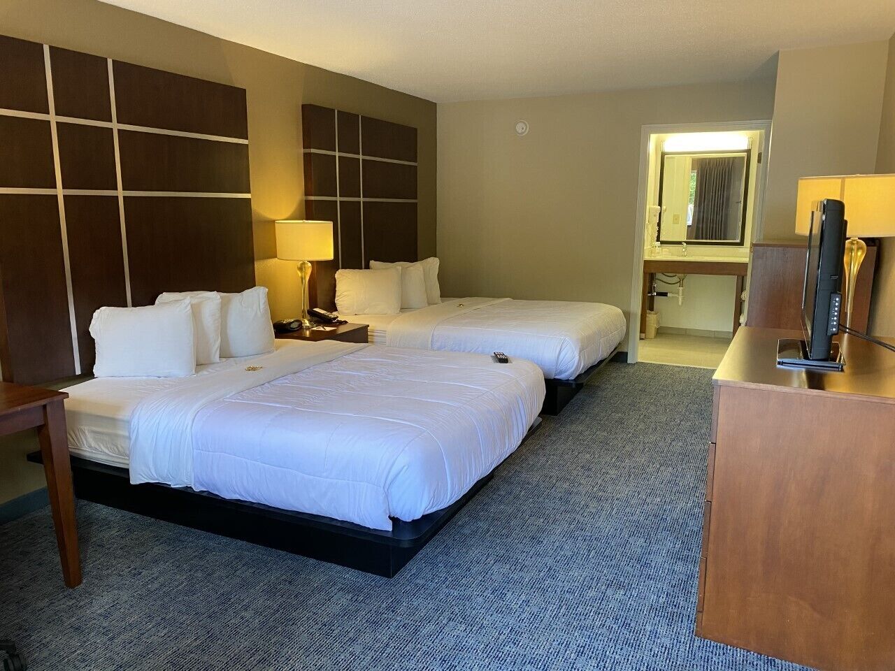A hotel room with two beds, a desk, a TV, and a bathroom visible through an open door.