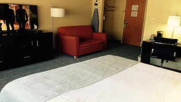 A hotel room with a large bed, a red sofa, a flat-screen TV, a desk with a lamp, and a door with a sign.