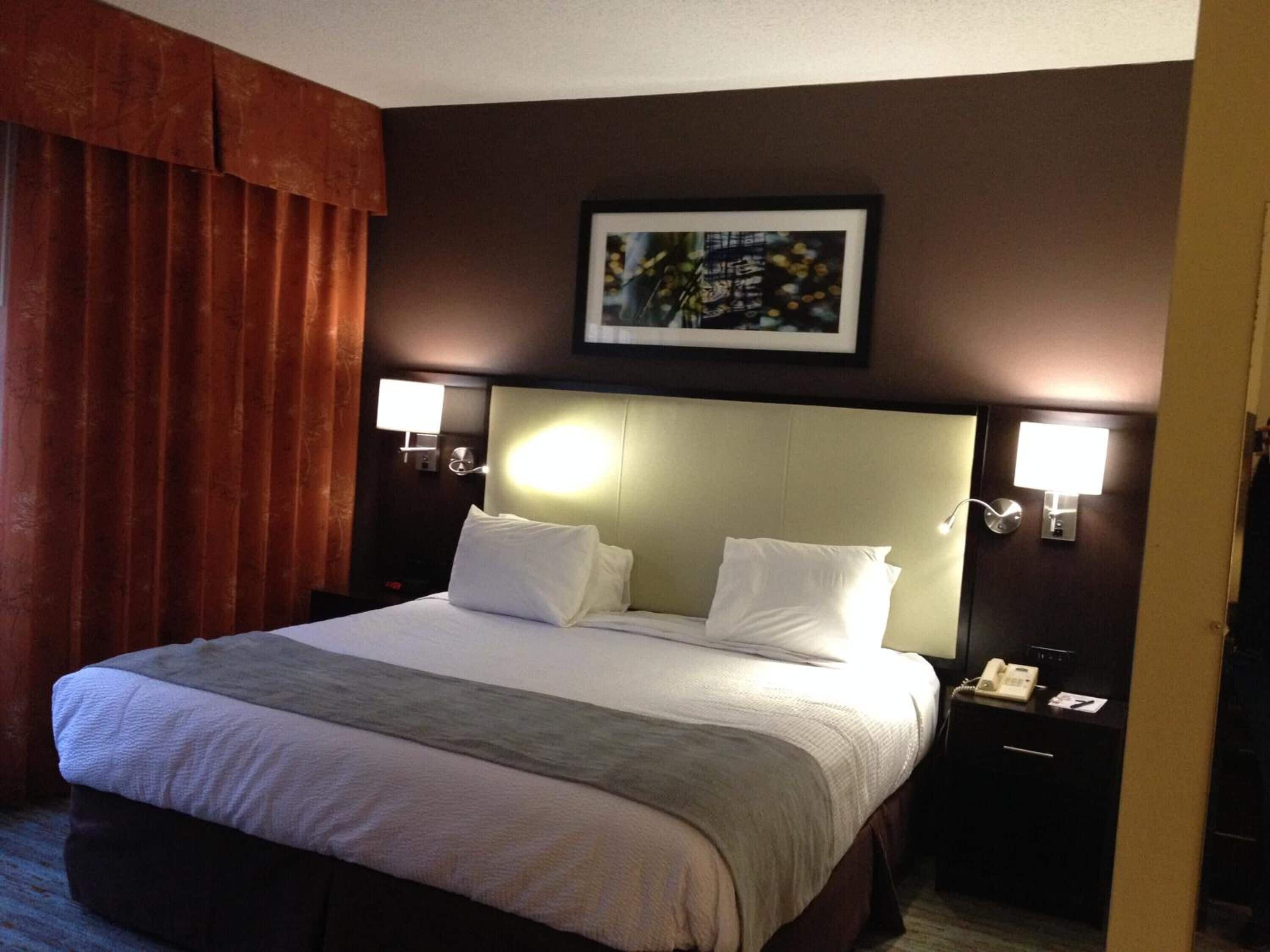 A hotel room with a large bed, two bedside tables, a nightstand, a framed picture on the wall, and a window with curtains.