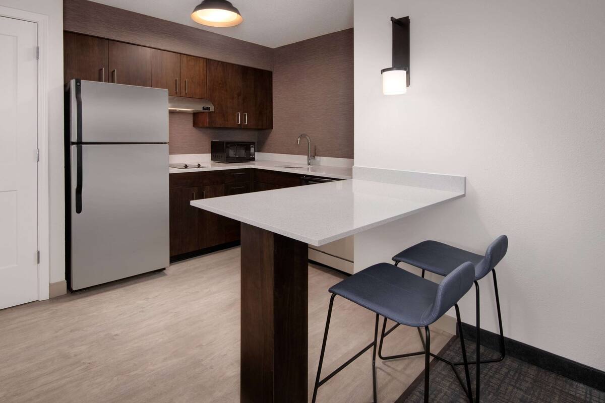 Une cuisine moderne avec un réfrigérateur en acier inoxydable, des meubles de cuisine en bois foncé, un plan de travail blanc et deux tabourets de bar.
