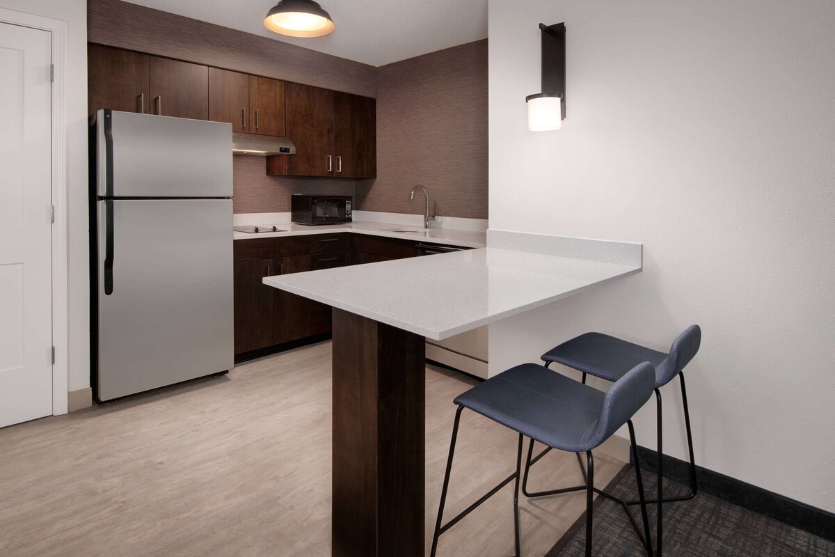 Una cocina moderna con un refrigerador de acero inoxidable, armarios de madera oscura, una encimera blanca y dos taburetes de bar.