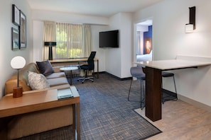 Ein Hotelzimmer mit Sofa, Schreibtisch, Sessel, Fernseher und einer Barzeile.