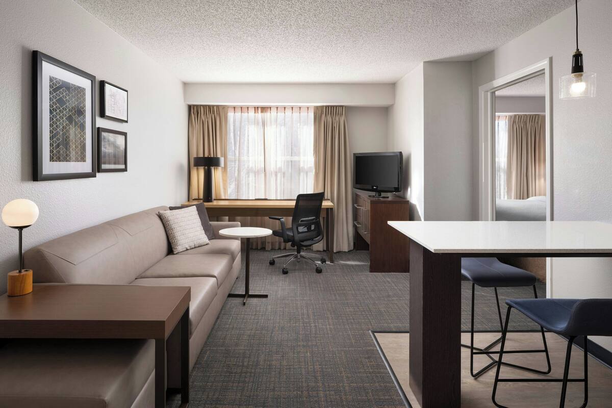 A modern hotel room with a sofa, a desk, a chair, a TV, and framed pictures on the wall.