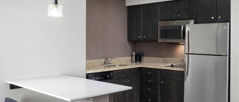 A modern kitchen with dark wood cabinets, a stainless steel refrigerator, a microwave, and a coffee maker. There is a white countertop and a small island with two chairs.