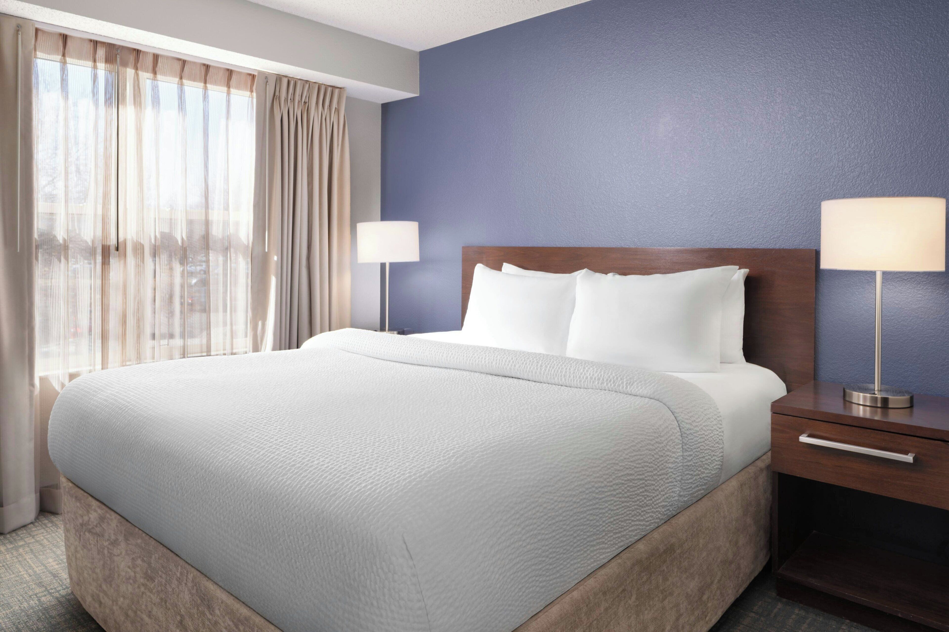 A hotel room with a large bed, two bedside tables with lamps, a window with sheer curtains, and a blue accent wall.