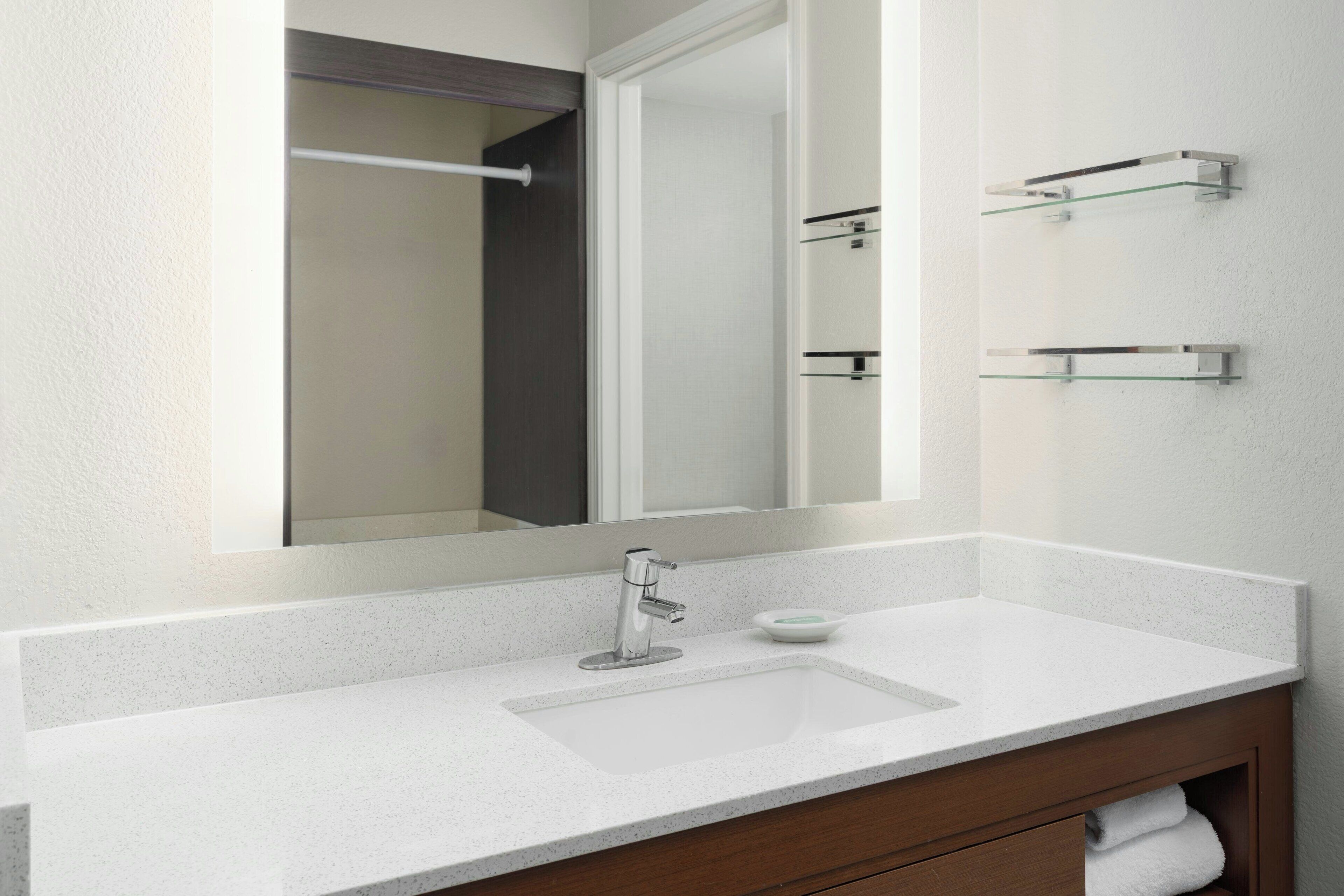 A modern bathroom with a white countertop, a single sink, a mirror, and two glass shelves.