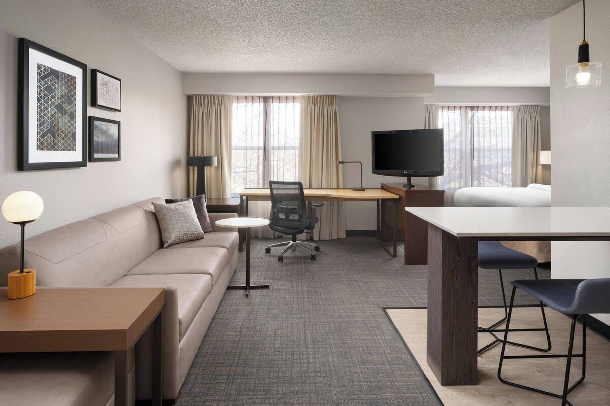 A modern hotel room with a sofa, a desk, a TV, and a dining table.